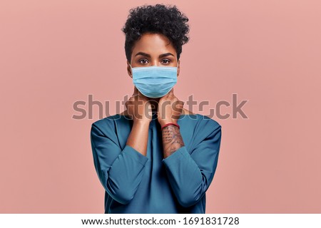 Gorgeous dark skinned young female with Afro hairstyle wearing protective mask against COVID virus, tired of stress and tension, looks confidently at the camera, poses against beige studio background