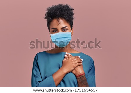 Portrait of kind hearted African American female paramedic wearing a respiratory mask from coronavirus disease, keeps hands on chest, shows her kindness and sympathy, on beige wall. COVID-19 epidemic