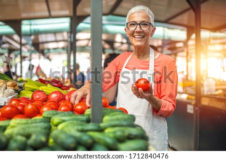 Woman tending an organic vegetable stall at a farmer's market and selling fresh vegetables. Female gardener selling organic crops and picking up a bountiful basket full of fresh produce. Only organic.