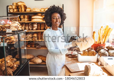 Smiling baker woman standing with fresh bread at bakery. Happy african woman standing in her bake shop and looking at camera. Satisfied baker with breads in background. Beautiful  woman at bakery shop