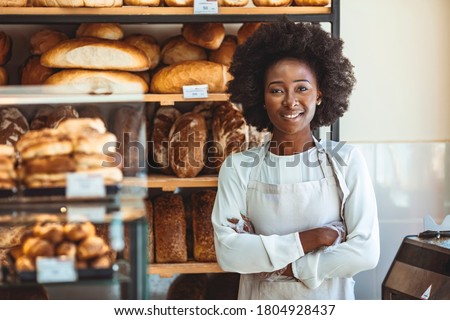 Portrait of positive African American young woman working in own Bakery shop, looking at camera with toothy smile. Pretty baker smiling at camera. Small Bakery shop owner standing in front of store