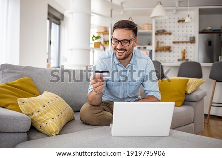 Happy cheerful smiling young adult man doing online shopping or e-shopping satisfied entrepreneur making online payment paying for service or goods self employed freelancer collecting fee