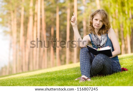 Cute smiling woman holding book  in the summer park and  points his finger to the up