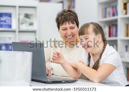 Smiling girl with down syndrome is uses a laptop with her teacher at library. Education for disabled children concept Foto d'archivio © 