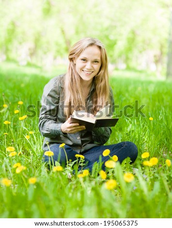 laughing girl sitting on grass with dandelions reading a book and looking at camera