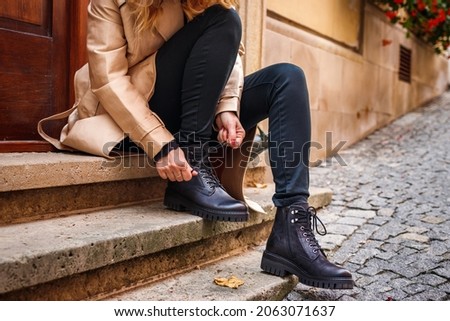Woman wearing trench coat sitting on staircase and tying shoelace on her ankle boot. Autumn fashion collection. Trendy black leather shoe. Street style Foto d'archivio © 