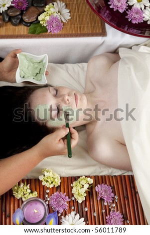 A young Caucasian woman lies on a massage table with a candle and flowers with a face mask being applied.