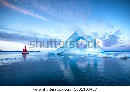 Sail boat with red sails cruising among massive ice bergs during dusk. Disko Bay, Greenland.