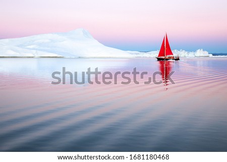 Sail boat with red sails cruising among ice bergs during dusk in pink and purple alpen glow light. Disko Bay, Greenland.