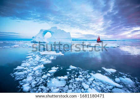 Sail boat with red sails cruising next to a pierced ice berg after sunset. Disko Bay, Greenland.