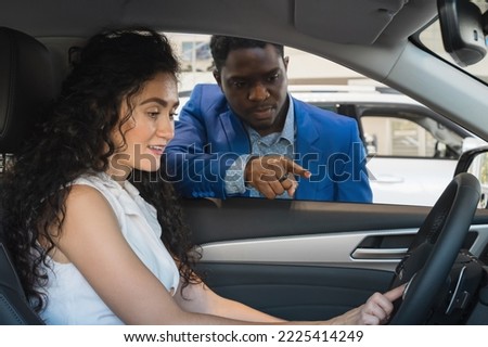 Black-haired lady driver with excited expression examines car listening to African American salesman presentation. Dealer talks about characteristics of vehicle Foto stock © 