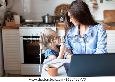 Working mom works from home office. Happy mother and daughter look to each other. Woman and cute child using laptop. Freelancer workplace in cozy kitchen. Female business, kindness, care. Lifestyle.