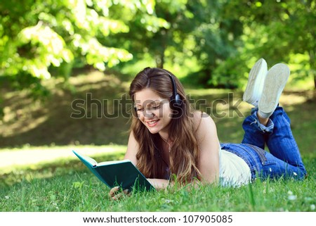 young woman listening music through headset and reading book in park