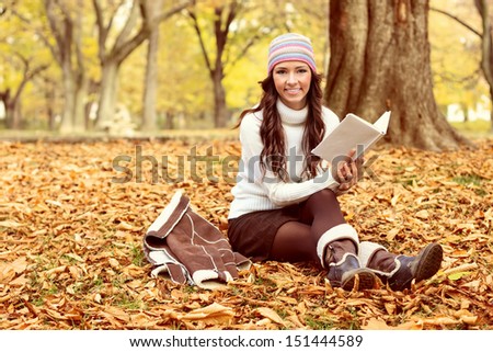 Young woman sitting on the autumn leaves and reading book in park