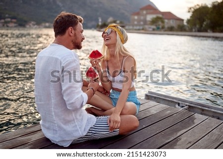 Photo of Couple having good time on date by sea siting on wooden dock, laughing and eating watermelon. Love, fun, togetherness concept.
