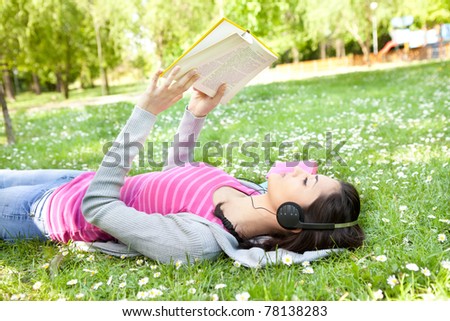 young woman listening music on the grass using headphones and reading book