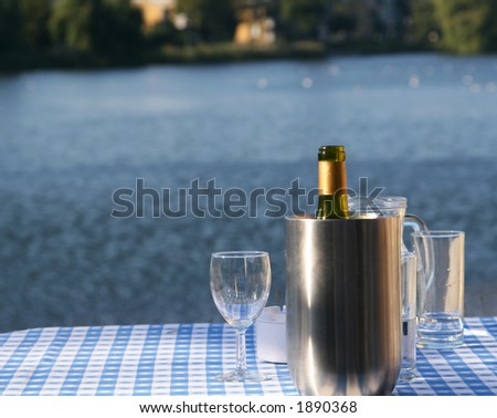 An open bottle of wine in a chilling bucket, glasses standing close by, set on a table with a lakescene in the background