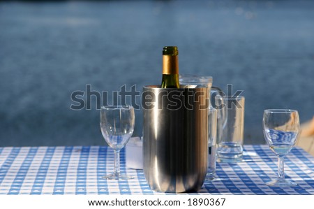 An open bottle of wine in a chilling bucket, glasses standing close by, set on a table with a lakescene in the background