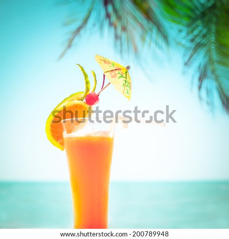 Tequila sunrise cocktail with fruits and umbrella decoration at tropical ocean beach with palm trees. Vintage style, hipster colors image with copy space for party invitation text
