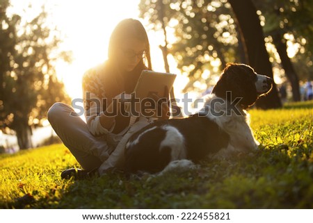 Girl Using Tablet In The Park On The Grass With The Dog At Sunset