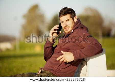 Handsome Sporty Man Sitting on The Bench Outdoor In Park And Talking On Mobile Phone While Making A Gesture With Hand