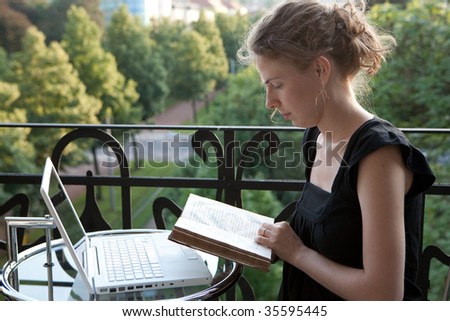 young woman writing on a white modern laptop computer and reading a book on a balcony in art nouveau