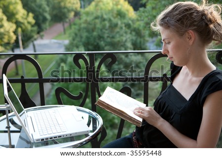 young woman writing on a white modern laptop computer and reading a book on a balcony in art nouveau