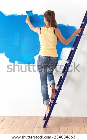 Beautiful young woman doing wall painting, standing on ladder