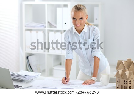 Portrait of female architect with blueprints at desk in office, isolated
