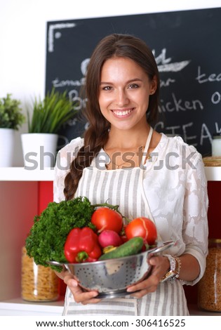 Smiling young woman holding vegetables standing in kitchen .