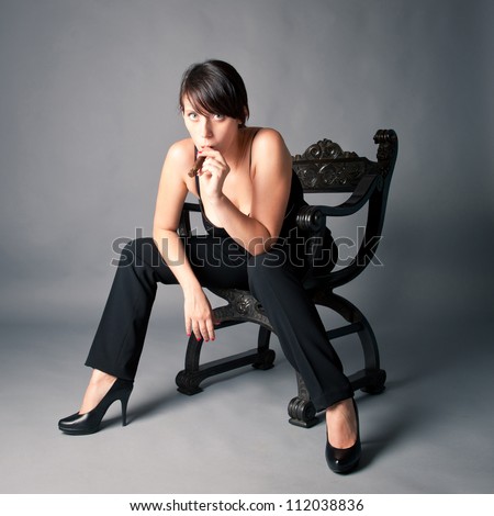 Woman smoking cigar sit on an old black chair. Studio fashion photo.