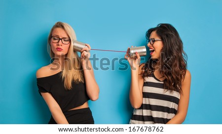 Blonde and brunette women talking with tin can telephone against blue background.