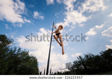 Attractive sexy woman pole dancer performing outdoors against blue cloudy sky.