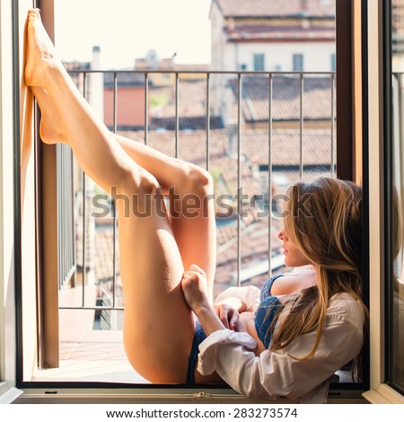 Sensual young blonde woman portrait wearing lingerie at the window in hotel room. Filtered image.