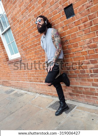 Young tattooed man portrait listening to music against brick wall in Shoreditch borough. London, UK. Hipster style.