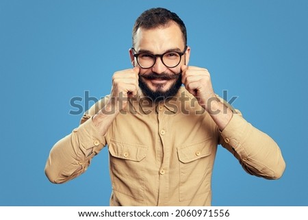 Hipster man with beard wearing eyeglasses adjusting his mustaches while standing against blue background ストックフォト © 