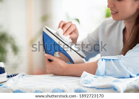 Close-up, woman wipes iron with soft cloth preparing to iron clothes on ironing board after washing at home Modern steam system, new household appliances. Caring for household appliances after work.