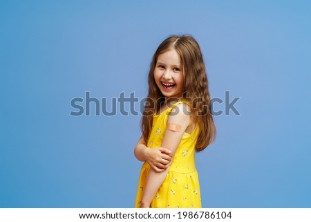 Happy Smiling Vaccinated Little Girl Shows Her Hand With A Patch After Being Injected With The Covid-19 Vaccine, Posing On A Blue Background. Advertising Of Vaccination Against Coronavirus