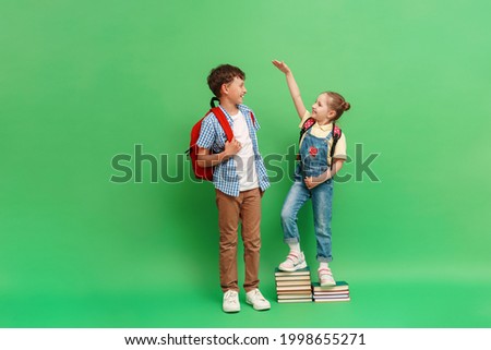 happy teenage schoolboy with a backpack, measuring the height of a little girl in loose clothes, standing on a stack of books on a green background. The concept of development and growth of education.