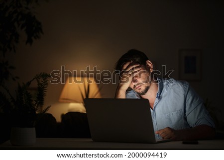 tired male businessman works at home at night on a laptop, works from a home office, studies online,. Internet addiction or a person who works late. a frightened man works late at his home office
