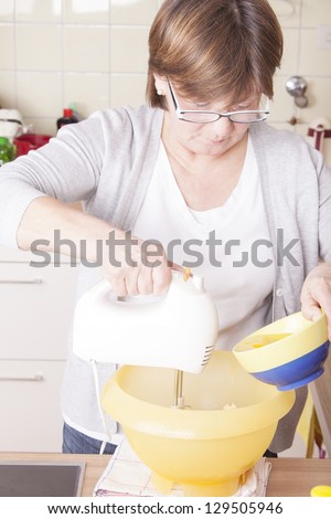 senior woman baking homemade chocolate cake, using a mixer and a bowl with eggs. baking chocolate/stracciatella cake in a glass jar.
