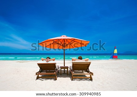 Couple on a tropical beach relax in the sun on deck chairs under a red umbrella.  Travel  background .