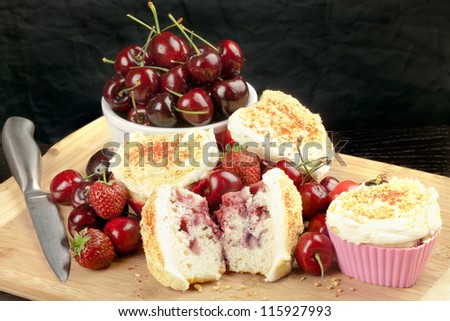 Close-up of a halved cream cheese iced strawberry cherry muffin surrounded by fruit and other muffins, topped with graham cracker crumbs.