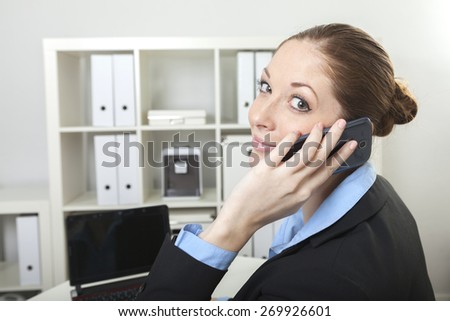 Friendly business lady sits at her desk while looking over her shoulder having a phone call on her cell phone