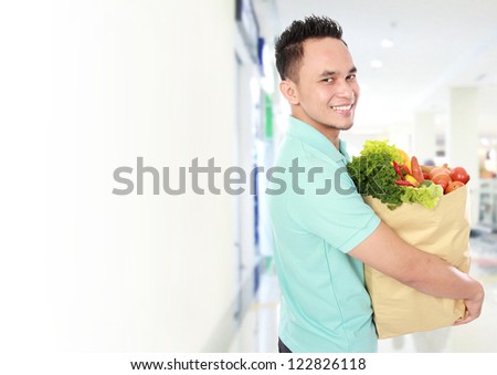 Portrait of smiling young man holding grocery bag full of groceries in supermarket