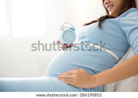 A portrait of a pregnant woman holding an alarm clock while sitting on the bed