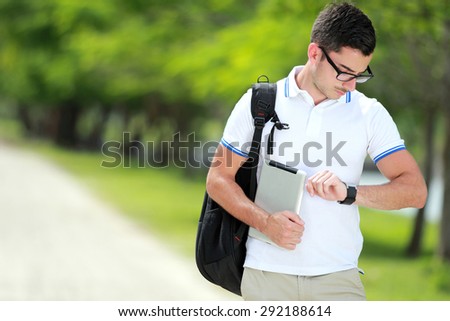 portrait of a handsome college student with backpack and tablet saw the time on his watch at college park with copy space