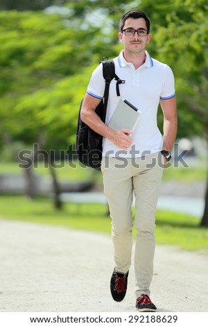 full length portrait of smiling handsome college student walking by at college park with backpack and holding a tablet