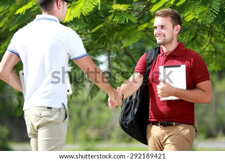 portrait of handsome college student with backpack smiling while shake hands with his friend at college park
