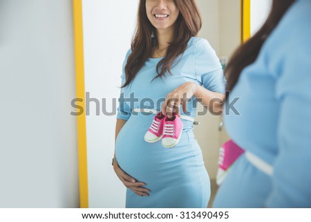 A portrait of Asian pregnant woman smiling in front of a mirror, holding a pair of baby shoes
